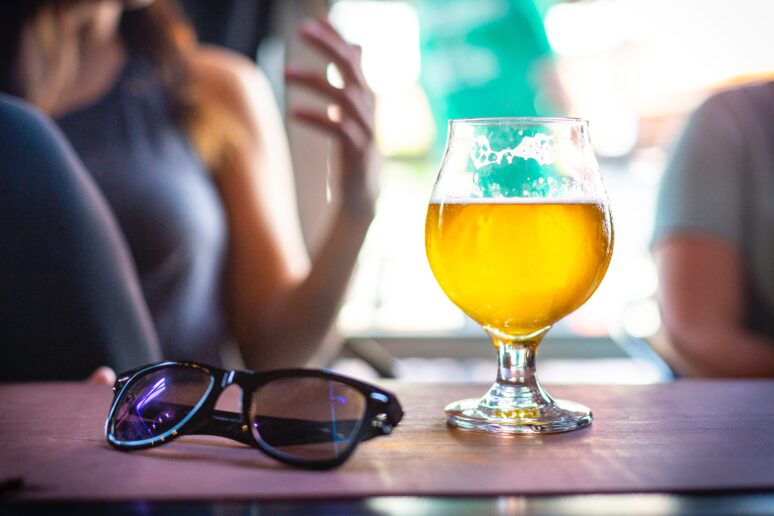 Alt-text: A glass of beer sits on top of a wooden tabletop next to a pair of sunglasses, while two people sit beside each other in the background. 